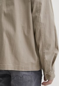 Beige shirt with loose fit, long sleeves, and buttoned cuff; fabric appears smooth with visible stitching. Gray jeans with distinctive pocket design.