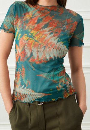 Vrouw die een doorschijnend teal shirt draagt met oranje en witte varenpatronen, gecombineerd met een olijfgroene broek met hoge taille en een dubbele ringriem.