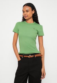 Zielony prążkowany t-shirt z krótkimi rękawami, okrągłym dekoltem i logo Lacoste; zestawiony z czarnymi, wysokim stanem spodniami oraz brązowym paskiem.
