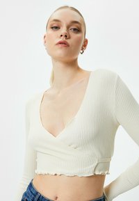 Cremefarvet ribbet langærmet crop top med dyb V-udskæring, med rå kant og tætsiddende silhuet. Matchet med højtaljede blå jeans.