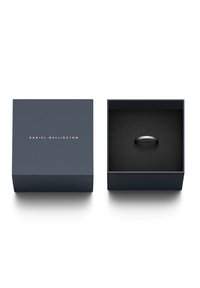 Anello nero esposto in un interno di velluto scuro di una scatola blu navy, caratterizzato da un design minimalista con il nome del marchio in oro sulla parte superiore.