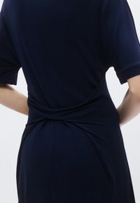 Rochie albastru închis cu mâneci scurte, având un detaliu răsucit în partea din spate și un material neted, ușor texturat. Design minimalist.