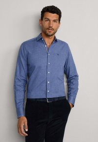Chemise à boutons en tissu bleu moyen avec une texture lisse. Comprend un col classique, des boutons blancs et un petit logo sur la poitrine.