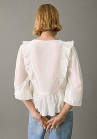 Bluză albă cu accente volănașe pe umeri, mâneci trei sferturi și talie peplum, asortată cu blugi ușor albaștri. Material moale, texturat.