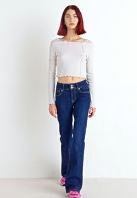 Haut court à manches longues en maille gris clair associé à un jean évasé bleu foncé. Boutons d'accent sur le jean. Le modèle porte des baskets roses.