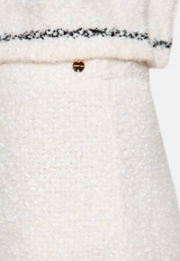 Cremfarbenes, strukturiertes Gewebe mit weichem, flauschigem Aussehen; verfügt über einen horizontalen schwarzen Streifen und ein kleines goldenes Logo-Akzent.