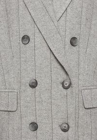 Manteau gris en chevrons à double boutonnage avec six boutons noirs et deux poches à rabat, mettant en valeur un tissu texturé et des revers structurés.