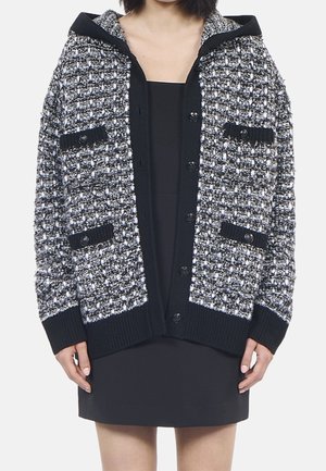 Femme portant un cardigan boutonné texturé noir et blanc avec quatre poches avant par-dessus une robe noire, visible des épaules jusqu'à mi-cuisse.