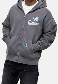 Sudadera gris con cremallera y bolsillos frontales, decorada con mariposas bordadas en rosa y verde. Fabricada en un tejido suave y texturizado.