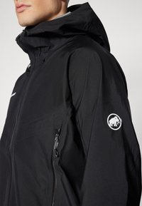 Veste imperméable noire avec un col haut et une capuche réglable. Équipée d'une poche latérale zippée et d'un patch logo blanc sur la manche.