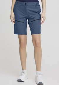 Shorts bleus avec une taille élastique, deux poches latérales zippées et une texture lisse, s'arrêtant au-dessus du genou. Portés avec des chaussures de sport blanches.
