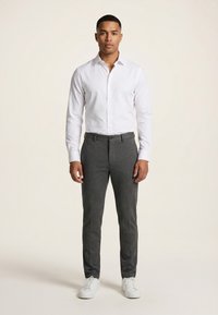 Homme debout avec une expression neutre, portant une chemise blanche à boutons, un pantalon gris foncé et des baskets blanches sur un fond uni.