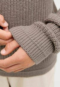 Gestrickter grauer Pullover mit einem strukturierten Rippenmuster. Nahaufnahme eines Ärmels, der eine lockere Passform und einen umgeschlagenen Rand zeigt, und das weiche Garn hervorhebt.