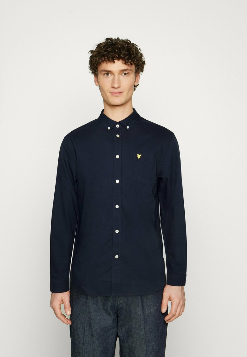 Camisa de botones de manga larga en color azul marino con cuello, que presenta un pequeño logo amarillo en el bolsillo izquierdo. Tejido suave y diseño ajustado.