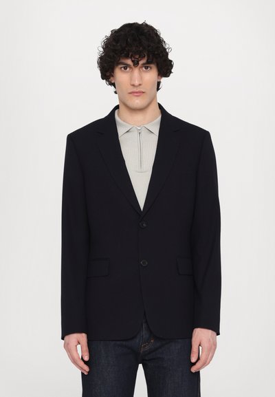 Navy blazer made of textured fabric with a single button closure, notched lapels, and two front pockets, worn over a light grey polo.
