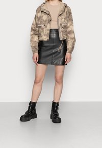 Veste de camouflage beige cintrée avec cordon de serrage, associée à une mini-jupe en cuir noir et des bottes de combat noires épaisses avec boucles.