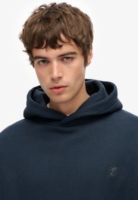 Sudadera con capucha de color azul marino, hecha de una tela suave con textura de canalé, que presenta un pequeño logo en el pecho y un ajuste relajado.