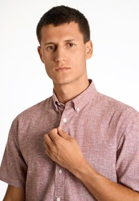 Jonge man met kort haar, gekleed in een roze kortetermijn overhemd met knopen, die de bovenste knoop dichtknopt, met een neutrale uitdrukking.