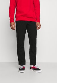 Pantalones negros de corte ajustado, combinados con una sudadera roja con bolsillo tipo canguro y puños acanalados, y zapatillas negras con detalles rojos.