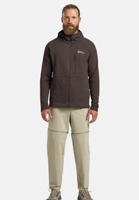Giacca marrone con zip, dotata di cappuccio e tasca frontale, abbinata a pantaloni chiari con zip, convertibili. Il materiale sembra sintetico e testurizzato.