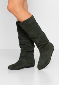 Tummanvihreät, polvimittaiset nahkabootsit, joissa on löysä muotoilu, pyöreä kärki ja mustat kumipohjat. Teksturoitu materiaali antaa pehmeän ilmeen.