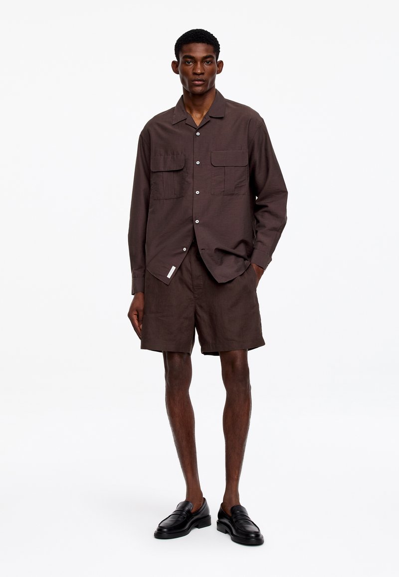 Homme portant une chemise à manches longues boutonnée en marron foncé avec deux poches poitrine, un short assorti et des chaussures noires sans lacets, debout les mains dans les poches.