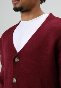 Cardigan tricoté bordeaux avec un col en V profond, doté de trois boutons ronds, en tissu doux et texturé. Porté sur une chemise blanche.
