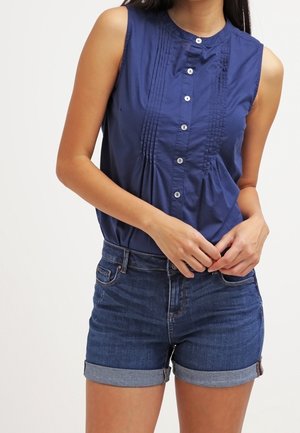 Blusa sin mangas azul marino con botones y pliegues, combinada con shorts de mezclilla oscuros remangados, en una persona de pie contra un fondo liso.