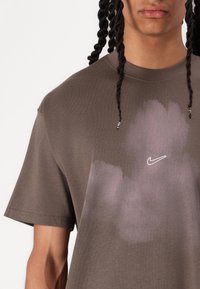 Hnědá bavlněná tričko s grafikou v purpurových odstínech a bílým logem Nike uprostřed. Krátké rukávy a kulatý výstřih.