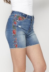 Denim shorts med blommig broderi på sidan, ljusblå färg, fransar i nederkanten och lätt slitage på framfickorna.
