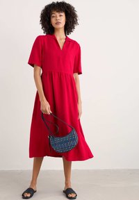 Rotes Kurzarmkleid mit Kragen, A-Linien-Form und geraffter Taille. Kombiniert mit einer blauen Musterhandtasche und schwarzen Rutschen.