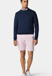 Marineblauw sweatshirt met een klein rood logo, gecombineerd met lichtroze shorts. Het model draagt ook een geruit overhemd met kraag en witte sneakers.