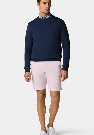 Marineblauw sweatshirt met een klein rood logo, gecombineerd met lichtroze shorts. Het model draagt ook een geruit overhemd met kraag en witte sneakers.