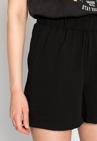 Shorts noirs à taille élastique avec une texture froissée, dotés de poches latérales et d'une coupe décontractée. Le matériau semble léger et respirant.