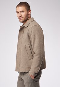 Beżowa wełniana kurtka z kołnierzykiem, przednimi kieszeniami i zapięciem na guziki. Gładka faktura, dopasowany krój i casualowy design.