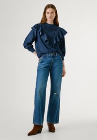 Top in denim con scollatura arricciata di colore blu scuro, abbinato a jeans a gamba larga blu con dettagli strappati e stivaletti con tacco marroni.