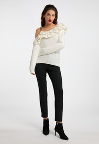 Witte off-the-shoulder trui met ruches en lange mouwen, gecombineerd met zwarte slim-fit broek en zwarte enkellaarsjes.