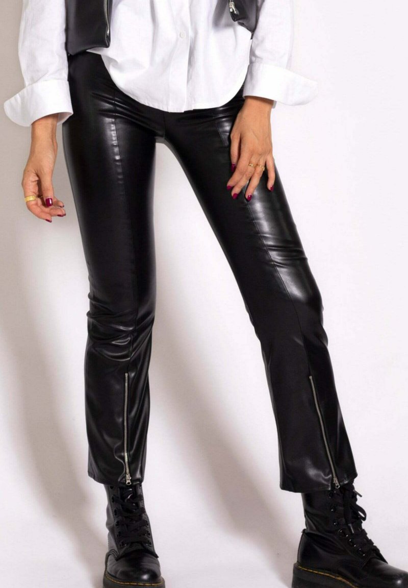 Pantalons en simili cuir noir à coupe ajustée, avec des zips sur les côtés aux chevilles, associés à une chemise blanche à boutons et des bottes à lacets noires.