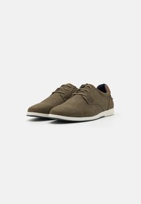 Chaussures en mesh marron à lacets avec une semelle blanche lisse, présentant une surface texturée et un petit accent en cuir au talon.