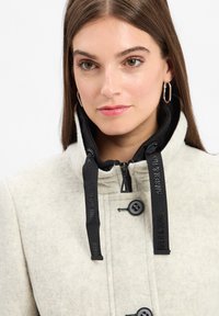 Junge Frau mit langen braunen Haaren, die einen hellen Wollmantel mit schwarzen Kordeln und Ohrhängern trägt, leicht nach rechts gewandt.