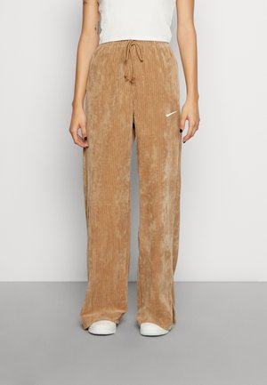 Person wearing loose-fitting light brown corduroy pants with white Nike logo and white sneakers, standing against a plain background.