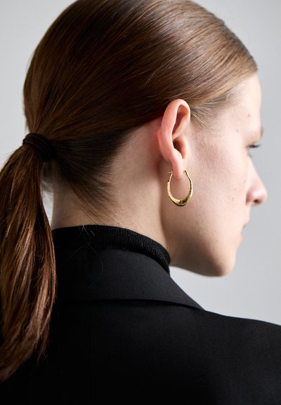 Pendiente de aro dorado con un diseño redondeado y ligeramente texturizado, llevado en la oreja izquierda frente a un fondo de cabello oscuro y una prenda negra.