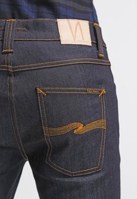 Tmavě modré džíny z denimu s koženou nášivkou na zadní straně s logem, oranžovým prošitím a výrazným vlnovým vzorem na zadní kapse.