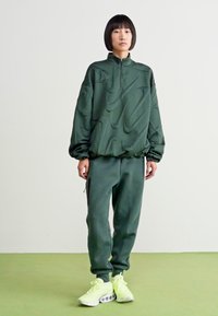 Πράσινος υφασμάτινος πουλόβερ με μισό φερμουάρ και ταιριαστές φόρμες. Και τα δύο διαθέτουν λεία υφή. Λαμπερά κίτρινα αθλητικά παπούτσια με ιδιαίτερο σχέδιο σόλας.