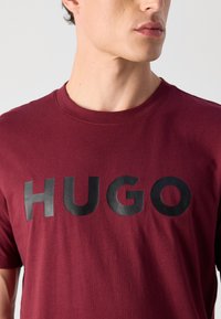 Fiatal férfi bordó pólót visel, amelyen nagy fekete betűkkel a "HUGO" felirat látható a mellkasán.