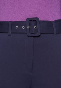 Mörkblå byxor med ett brett bälte med fyrkantig spänne, fem ögla hål och slät, strukturerad väv.