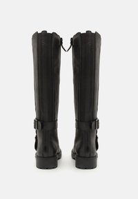 Botas de cuero negras hasta la rodilla con costuras verticales, detalles de hebillas en el tobillo y una suela de textura gruesa.