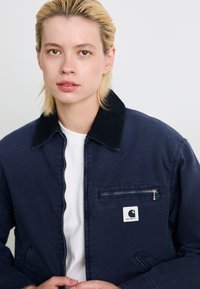 Young person with short blonde hair wearing navy blue Carhartt jacket over white shirt, facing forward against plain light background.
