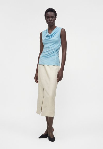 Γυναίκα που φοράει ένα αμάνικο ανοιχτό γαλάζιο τοπ με drape, μπέζ μίντι φούστα με μπροστινή σχισμή και μαύρα μυτερά παπούτσια, στέκεται μπροστά σε λευκό φόντο.