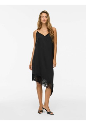 Femme portant une robe noire sans manches avec un ourlet à franges et des sandales noires à talons hauts, debout sur un fond blanc.